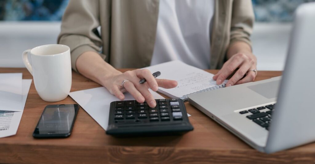 A person using a calculator and laptop while taking notes at a wooden desk.