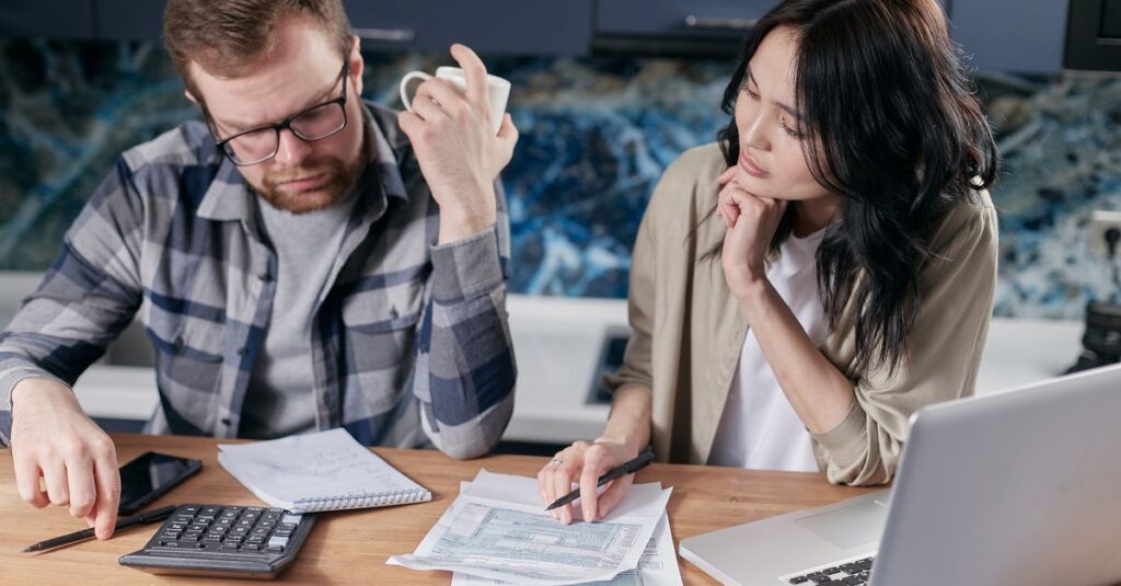 A couple reviews bills and documents on a laptop while discussing household finances.