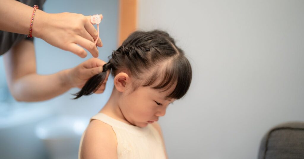 A young girl with braided hair looks at a smartphone indoors while receiving hair styling assistance.