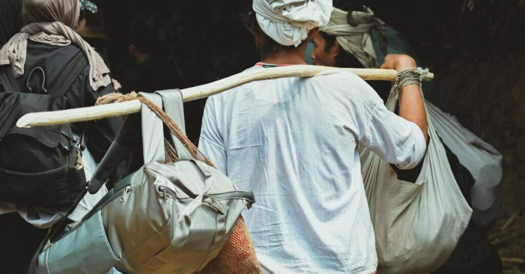A traveler carrying multiple bags using a wooden pole, going through a rural setting.