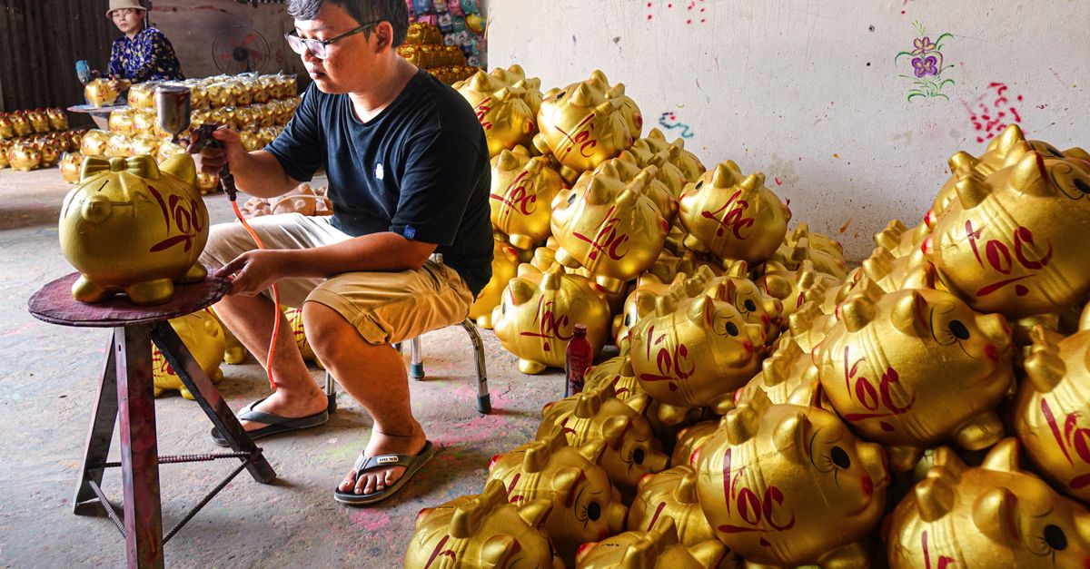 A craftsman expertly paints golden piggy banks in a workshop, surrounded by a large inventory.