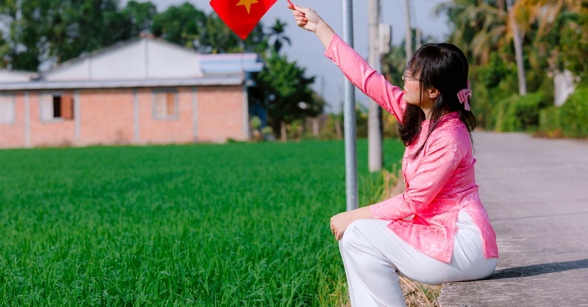 A woman in traditional attire waves the Vietnamese flag while sitting by a rural road.