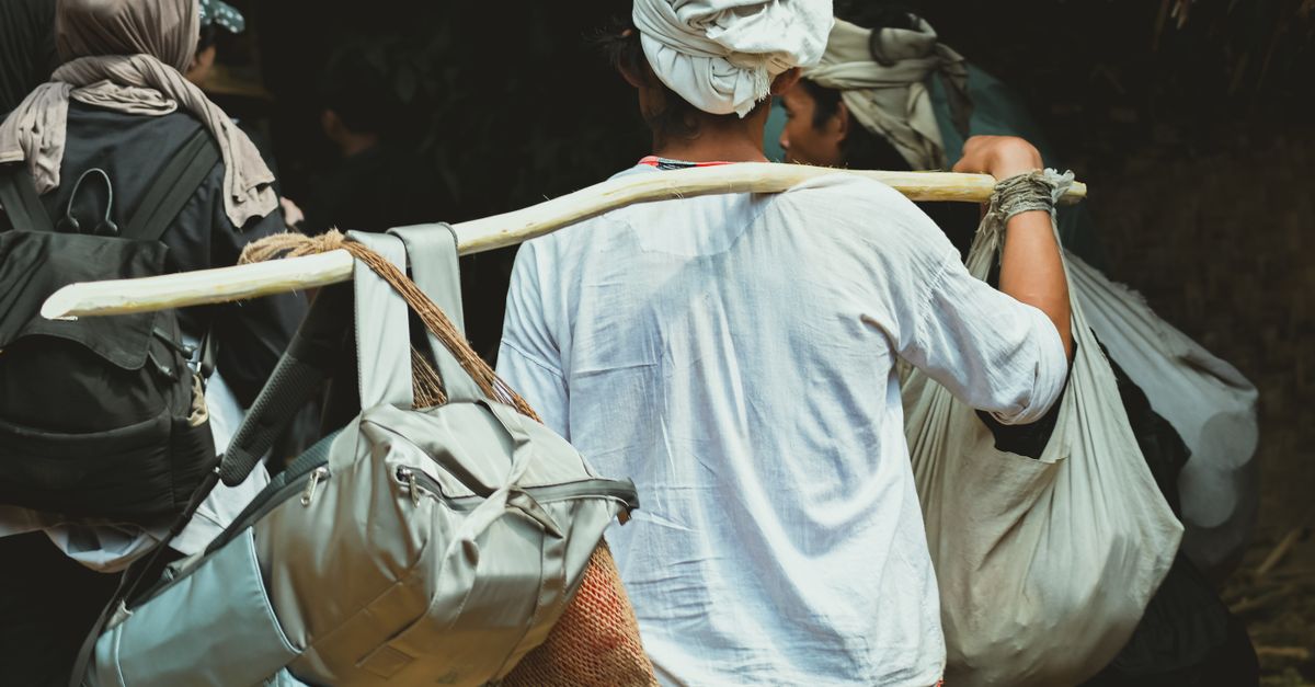 A traveler carrying multiple bags using a wooden pole, going through a rural setting.