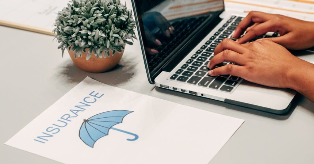 Hands typing on a laptop at a desk with an insurance paper and plant, suggesting a work environment.