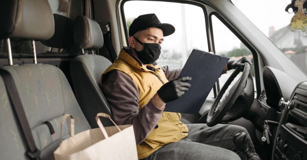 Masked delivery driver reading clipboard in a van during the day.