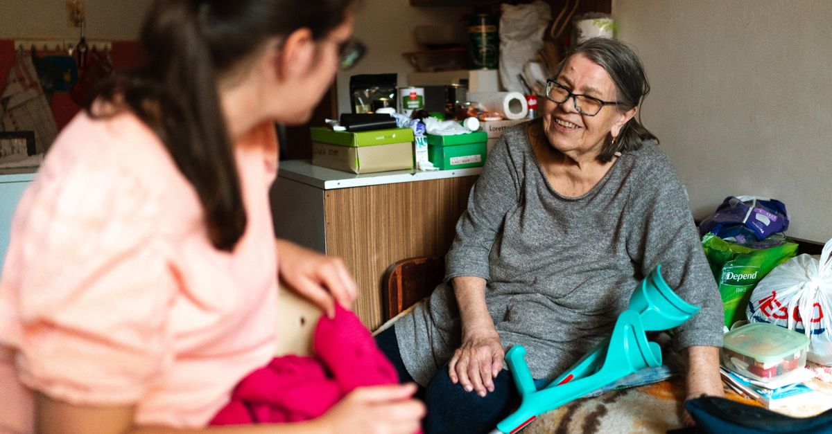 A caregiver helps a senior woman with a crutch in a cozy Prague home setting.