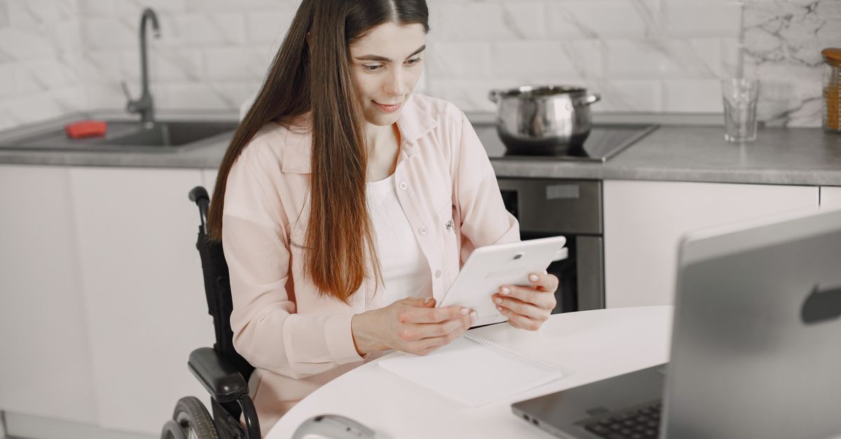 Woman in wheelchair engaged with tablet at kitchen table, creating an inclusive work environment.