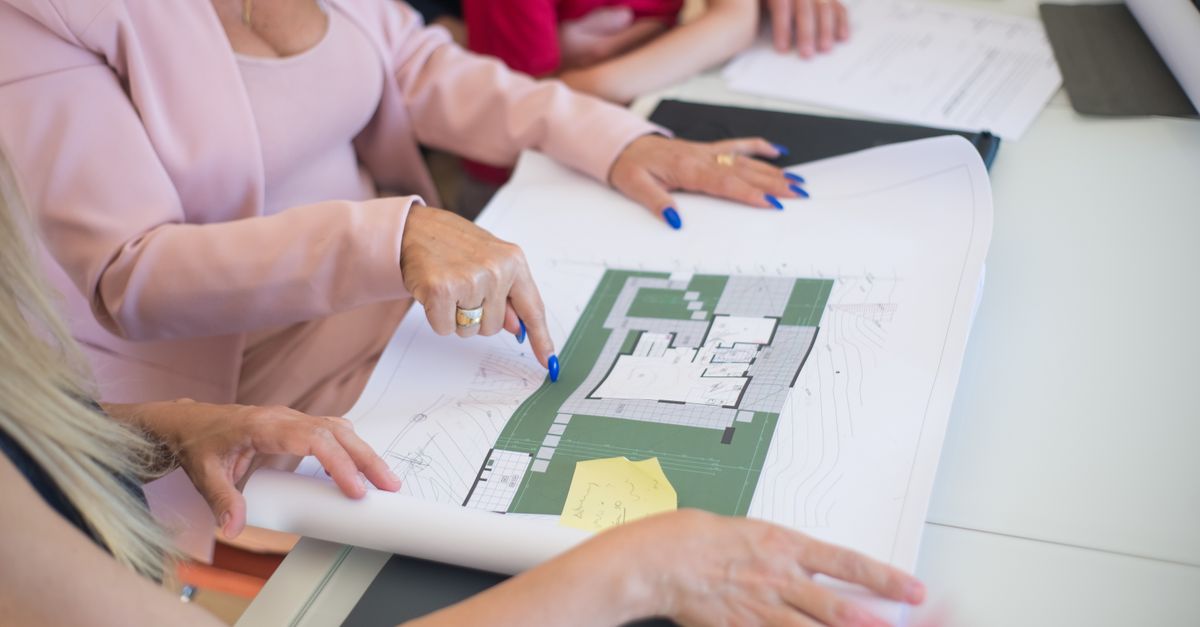 A real estate agent in a pink blazer shows floor plans to clients, discussing property details.